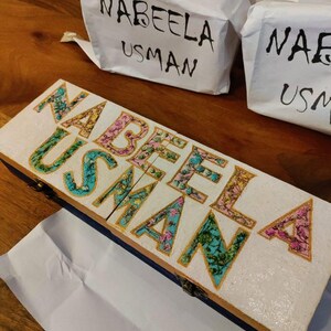 May include: A white wooden box with the name "NABEELA USMAN" spelled out in colourful letters with a floral pattern. The box is closed and has a gold trim.