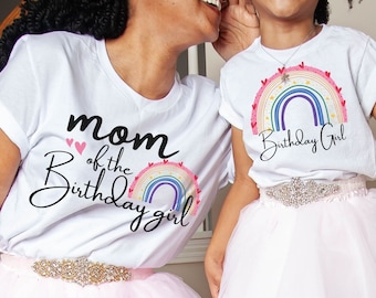 May include: Two white t-shirts with rainbow graphics and text that reads "Mom of the Birthday Girl" and "Birthday Girl". Both wear pink tulle skirts and jeweled belts. A mother and daughter are smiling.