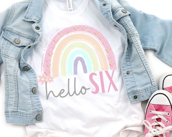 May include: A white t-shirt with a rainbow design and the text "hello six" in pink and gray. The rainbow is made of pastel colors and has floral accents. The shirt is paired with a denim jacket and pink sneakers.