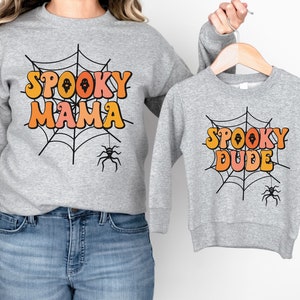 May include: Two heather gray crewneck sweatshirts featuring a black spiderweb graphic with a hanging spider. The larger sweatshirt displays the text SPOOKY MAMA in orange and pink, while the smaller sweatshirt displays SPOOKY DUDE in the same colors.