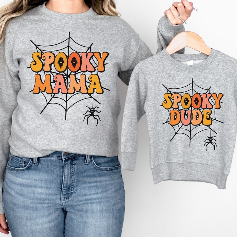 May include: Two heather gray crewneck sweatshirts featuring a black spiderweb graphic with a hanging spider. The larger sweatshirt displays the text SPOOKY MAMA in orange and pink, while the smaller sweatshirt displays SPOOKY DUDE in the same colors.
