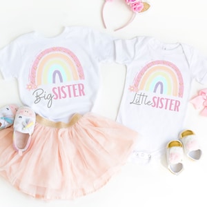 May include: Two white t-shirts with a rainbow and floral design. One shirt says "Big Sister" and the other says "Little Sister".