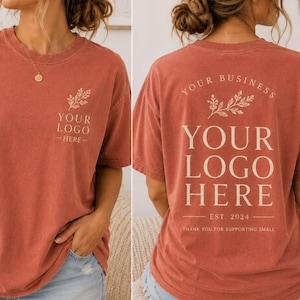 May include: A brick red t-shirt featuring a small cream-colored floral graphic and the text YOUR LOGO HERE on the front left chest. The back displays a larger version of the same floral graphic with the text YOUR BUSINESS, YOUR LOGO HERE, EST. 2024, and THANK YOU FOR SUPPORTING SMALL.