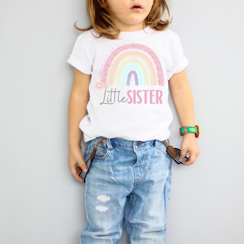 May include: White t-shirt with a pastel rainbow design and the text "Little Sister". The rainbow features pink, orange, yellow, green, and purple arches. The shirt is paired with blue jeans.