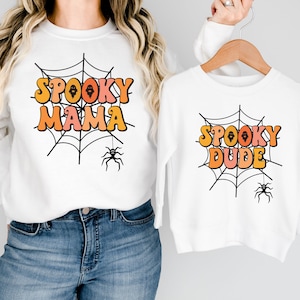 May include: Two white long-sleeve sweatshirts featuring a black spiderweb graphic with a hanging spider. One sweatshirt displays the orange and pink text "SPOOKY MAMA" and the other displays the orange and pink text "SPOOKY DUDE" in a retro font.