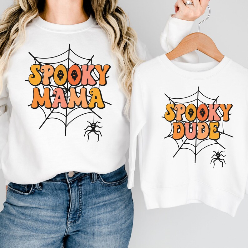 May include: Two white long-sleeve sweatshirts featuring a black spiderweb graphic with a hanging spider. One sweatshirt displays the orange and pink text "SPOOKY MAMA" and the other displays the orange and pink text "SPOOKY DUDE" in a retro font.