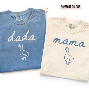 May include: Two Comfort Colors t-shirts. The blue shirt features the white text 'dada' above a white line-art goose. The cream shirt features the blue text 'mama' above a blue line-art goose. Both shirts have a crew neck and a visible brand label.