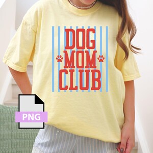 May include: Yellow t-shirt with the text "DOG MOM CLUB" in red, with blue vertical stripes and paw prints. The shirt is a light yellow color.