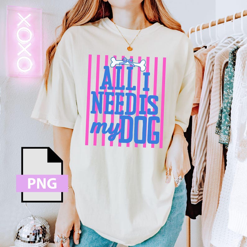 May include: Cream-colored t-shirt with the text "ALL I NEED IS my DOG" in blue, set against pink vertical stripes. A dog bone graphic is above the text. The shirt is part of a casual wear collection.