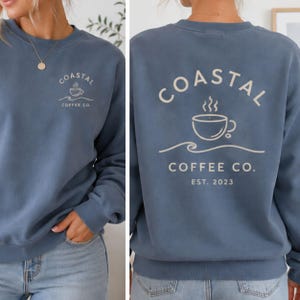 May include: A blue crewneck sweatshirt featuring a small graphic on the front left chest and a large graphic on the back. Both graphics display a coffee cup icon above a wave line with the text Coastal Coffee Co. The back includes Est. 2023.