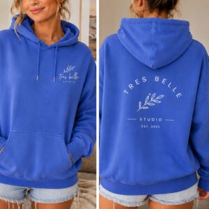 May include: A bright blue hooded sweatshirt featuring a front kangaroo pocket and a white graphic on the chest that reads "tres belle STUDIO" with a leaf sprig. The back displays the same text in a circular arc above a leaf sprig and "EST. 2023".