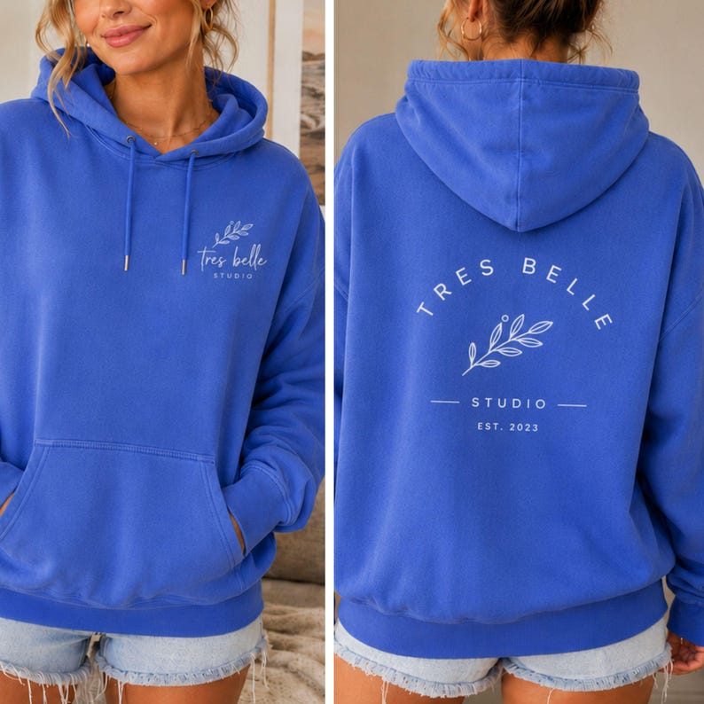 May include: A bright blue hooded sweatshirt featuring a front kangaroo pocket and a white graphic on the chest that reads "tres belle STUDIO" with a leaf sprig. The back displays the same text in a circular arc above a leaf sprig and "EST. 2023".