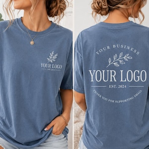 May include: A blue short-sleeve t-shirt featuring a white graphic on the front left chest with a leaf sprig and the text YOUR LOGO EST. 2024. The back displays a larger version of the same design with the text YOUR BUSINESS and THANK YOU FOR SUPPORTING SMALL.