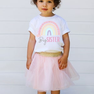 May include: A young child wearing a white t-shirt with a rainbow graphic and the words "Big Sister" in gray and pink. The child is also wearing a pink tulle skirt with a gold waistband.