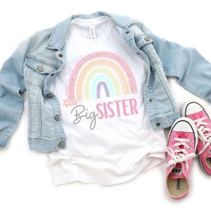 May include: White t-shirt with a pastel rainbow graphic and the text "Big Sister" in pink and gray. A light blue denim jacket and pink sneakers with white laces are also in the image.