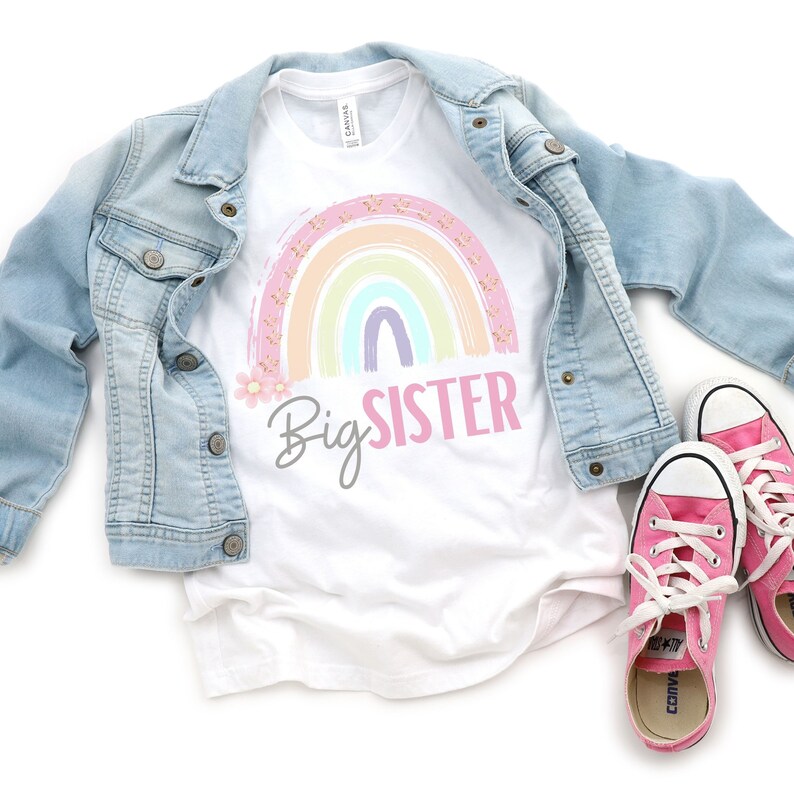 May include: White t-shirt with a pastel rainbow graphic and the text "Big Sister" in pink and gray. A light blue denim jacket and pink sneakers with white laces are also in the image.