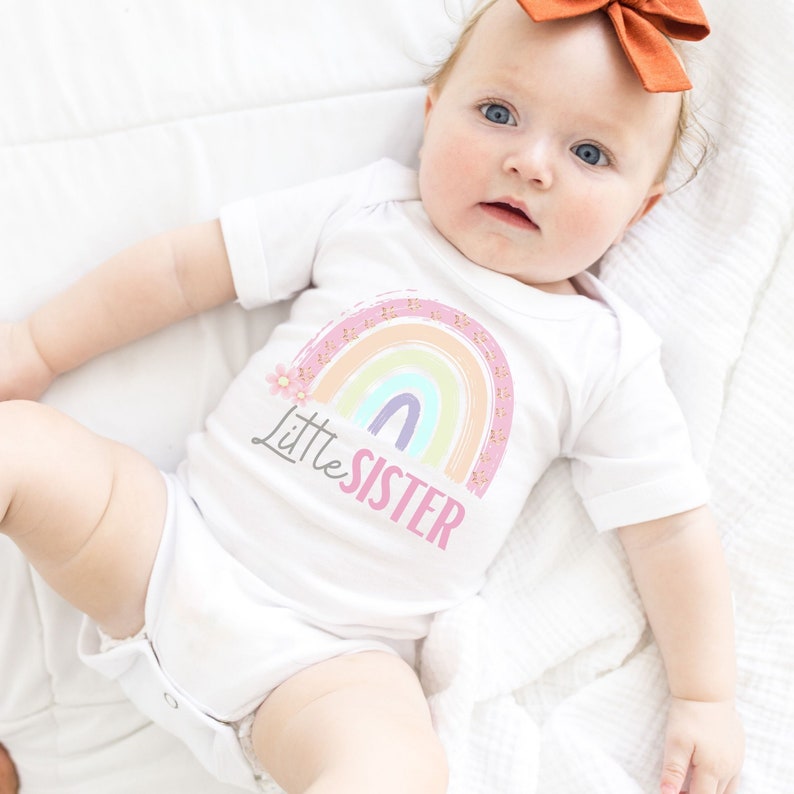 May include: A white baby onesie with a pastel rainbow design and the words "Little Sister" printed on the front. The rainbow includes pink, orange, yellow, green, and purple colors. A small pink flower is next to the rainbow.