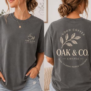 May include: A charcoal gray short-sleeve t-shirt featuring a small coffee cup graphic and the text OAK & CO. COFFEE on the front left chest. The back displays a large leaf illustration with the text GOOD COFFEE OAK & CO. COFFEE MADE TO BRING PEOPLE TOGETHER.