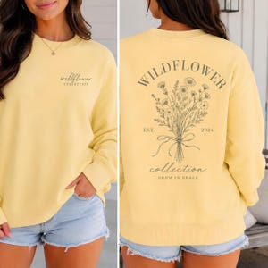 May include: A pale yellow crewneck sweatshirt featuring the text wildflower COLLECTION on the front left chest. The back displays a large botanical illustration of a wildflower bouquet with the text WILDFLOWER COLLECTION EST. 2024 GROW IN GRACE.
