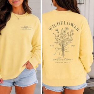 May include: A pale yellow crewneck sweatshirt featuring a small dark gray wildflower collection logo on the front chest and a large botanical bouquet illustration on the back with the text Wildflower Collection Est 2024 Grow In Grace.