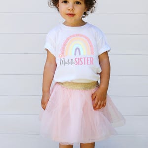 May include: A white t-shirt with a rainbow design and the text "Middle Sister" paired with a pink tulle skirt and a gold waistband. The outfit is for a young child.