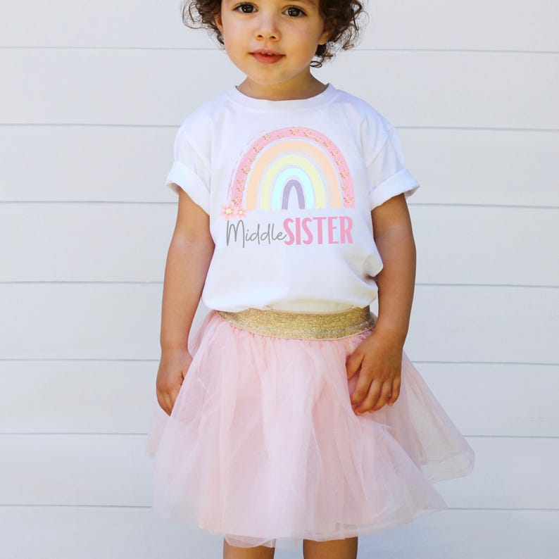 May include: A white t-shirt with a rainbow design and the text "Middle Sister" paired with a pink tulle skirt and a gold waistband. The outfit is for a young child.