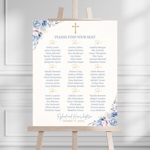 May include: A cream-colored seating chart on an easel, adorned with floral accents in blue and pink. The chart features a gold cross and the text "PLEASE FIND YOUR SEAT." Names are listed under table numbers, with the text "Ryland and Knox's baptism October 11, 2025."