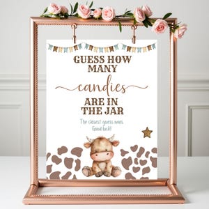 May include: A rose gold frame holds a sign that reads "GUESS HOW MANY candies ARE IN THE JAR." The sign features a cartoon cow, brown cow spots, and a star. The sign is decorated with small pennant banners.
