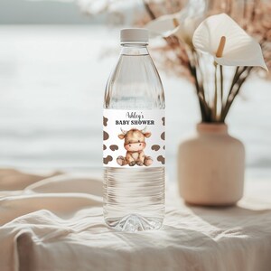 May include: Clear plastic water bottle with a white label featuring the text "Ashley's BABY SHOWER" and a watercolour illustration of a brown and tan calf. The label also has brown cow spots.
