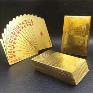 Puede incluir: Un juego de cartas doradas abanicadas, mostrando el As de corazones. Las cartas tienen símbolos y números rojos. También se ven una caja de cartas dorada y una pila de cartas, todas con un acabado metálico.