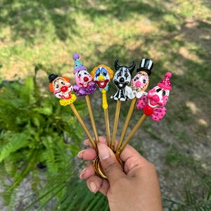 May include: A hand holds a set of six clown-themed decorative sticks. Each stick features a different clown face with colorful details, including hats, makeup, and expressions. The sticks are gold-colored and appear to be attached to a gold ring.