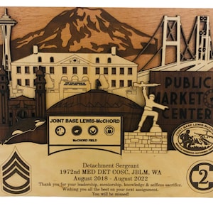 May include: A wooden plaque featuring laser-cut depictions of Seattle landmarks, including the Space Needle, a mountain, and a bridge. The plaque also includes text that reads "Joint Base Lewis-McChord" and "Public Market Center". The plaque is a commemorative item.
