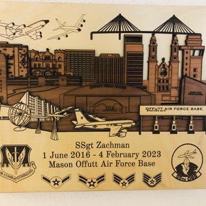 May include: A wooden plaque with a laser-cut design of a cityscape, two airplanes, and a military base. The plaque includes the text "Offutt Air Force Base", "SSgt Zachman", "1 June 2016 - 4 February 2023", and "Air Combat Command".