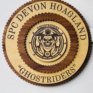 May include: A wooden, circular plaque with the text "SPC DEVON HOAGLAND" and "GHOSTRIDERS". The center features a skull wearing a hat. The plaque is made of light and dark brown wood.