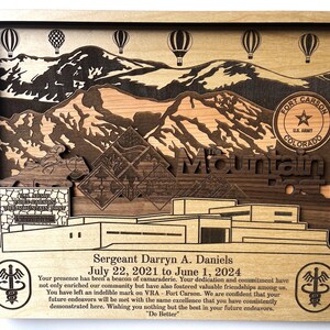 May include: A wooden plaque with a laser-cut design of mountains, hot air balloons, and a building. The plaque has the text "U.S. Army Fort Carson The Mountain Post" and "Sergeant Darryn A. Daniels July 22, 2021 to June 1, 2024" on it.