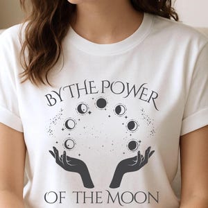A woman wearing a white t-shirt with a graphic design featuring a hand holding a crescent moon and the text "By the Power of the Moon".