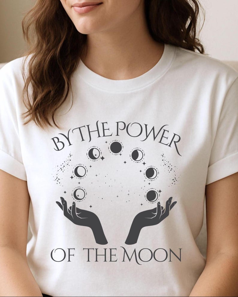 A woman wearing a white t-shirt with a graphic design featuring a hand holding a crescent moon and the text "By the Power of the Moon".