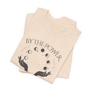 a folded beige t-shirt with a graphic design featuring a pair of hands holding a crescent moon and the text "BY THE POWER" above it.