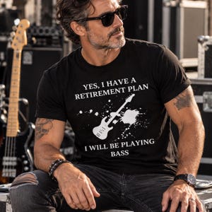 May include: A black short-sleeved t-shirt featuring a white graphic of a bass guitar with paint splatter effects and the text: YES, I HAVE A RETIREMENT PLAN I WILL BE PLAYING BASS. The shirt has a crew neckline and a standard fit.