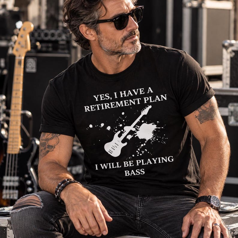 May include: A black short-sleeved t-shirt featuring a white graphic of a bass guitar with paint splatter effects and the text: YES, I HAVE A RETIREMENT PLAN I WILL BE PLAYING BASS. The shirt has a crew neckline and a standard fit.