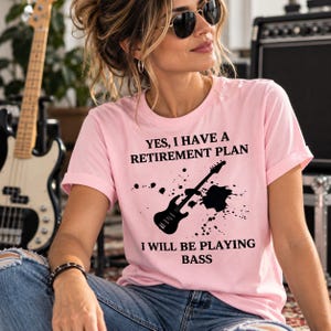 May include: A light pink short-sleeved t-shirt featuring a black graphic of a bass guitar with ink splatter effects. The text reads: YES, I HAVE A RETIREMENT PLAN I WILL BE PLAYING BASS. The shirt has a crew neckline and a casual fit.