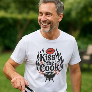 A smiling man wearing a white t-shirt with a graphic design that says "Kiss the Cook".