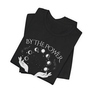 a black t-shirt with a white graphic design on it. The design features a pair of hands holding a bunch of planets, with the phrase "BY THE POWER" written above the hands.