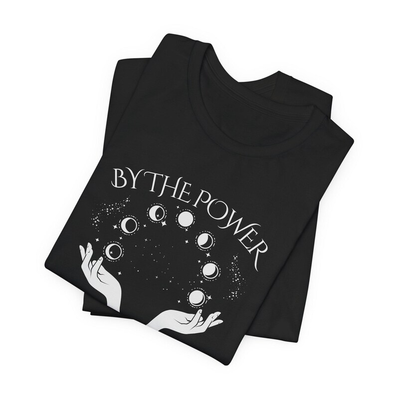 a black t-shirt with a white graphic design on it. The design features a pair of hands holding a bunch of planets, with the phrase "BY THE POWER" written above the hands.