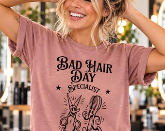 Może przedstawiać: Koszulka z krótkim rękawem w kolorze przybrudzonego różu z czarnym napisem graficznym Bad Hair Day Specialist. Poniżej tekstu znajdują się dwie postacie z kreskówek, jedna w kształcie nożyczek, a druga grzebienia, obie z narysowanymi twarzami.
