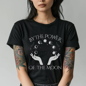 A woman wearing a black t-shirt with a graphic design featuring a hand holding a crescent moon and the text "BY THE POWER OF THE MOON".