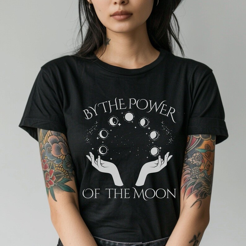 A woman wearing a black t-shirt with a graphic design featuring a hand holding a crescent moon and the text "BY THE POWER OF THE MOON".