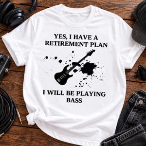 May include: A white short-sleeved t-shirt featuring a black graphic of a bass guitar surrounded by ink splatters. The text reads: YES, I HAVE A RETIREMENT PLAN I WILL BE PLAYING BASS. The design is centered on the front of the garment.