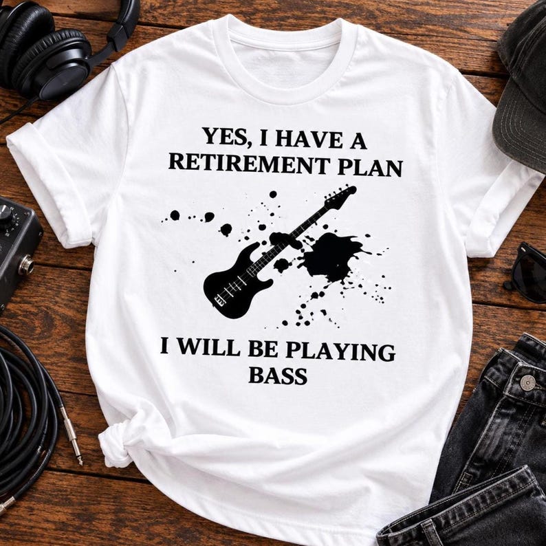 May include: A white short-sleeved t-shirt featuring a black graphic of a bass guitar surrounded by ink splatters. The text reads: YES, I HAVE A RETIREMENT PLAN I WILL BE PLAYING BASS. The design is centered on the front of the garment.