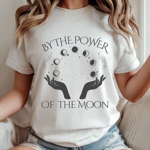 A woman wearing a white t-shirt with the text "BY THE POWER OF THE MOON" printed on it.